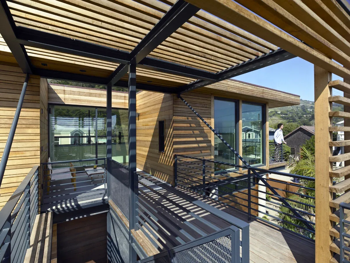 Elevated walkway with wood slat pergola and metal railing connects different parts of the wood-sided house.