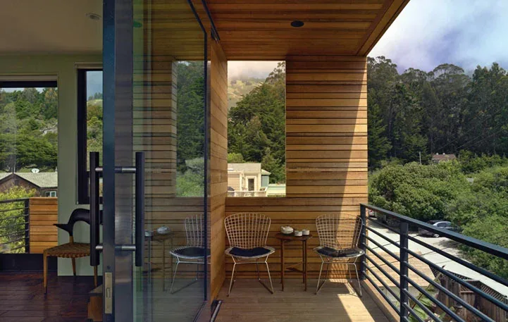 The balcony features wood siding and ceiling with metal railing and wire chairs, offering views of greenery and hills.