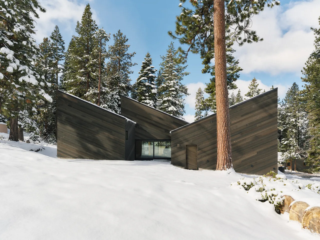 A modern cabin with staggered shed roofs and dark wood siding stands in a snowy forest landscape.