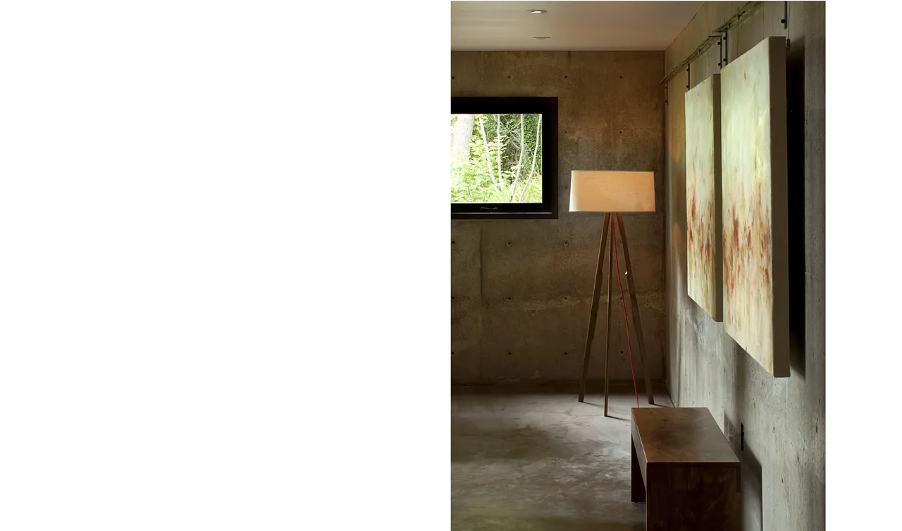 An interior view of a minimalist room with exposed concrete walls, a small window, wooden bench, tripod lamp, and abstract wall art.