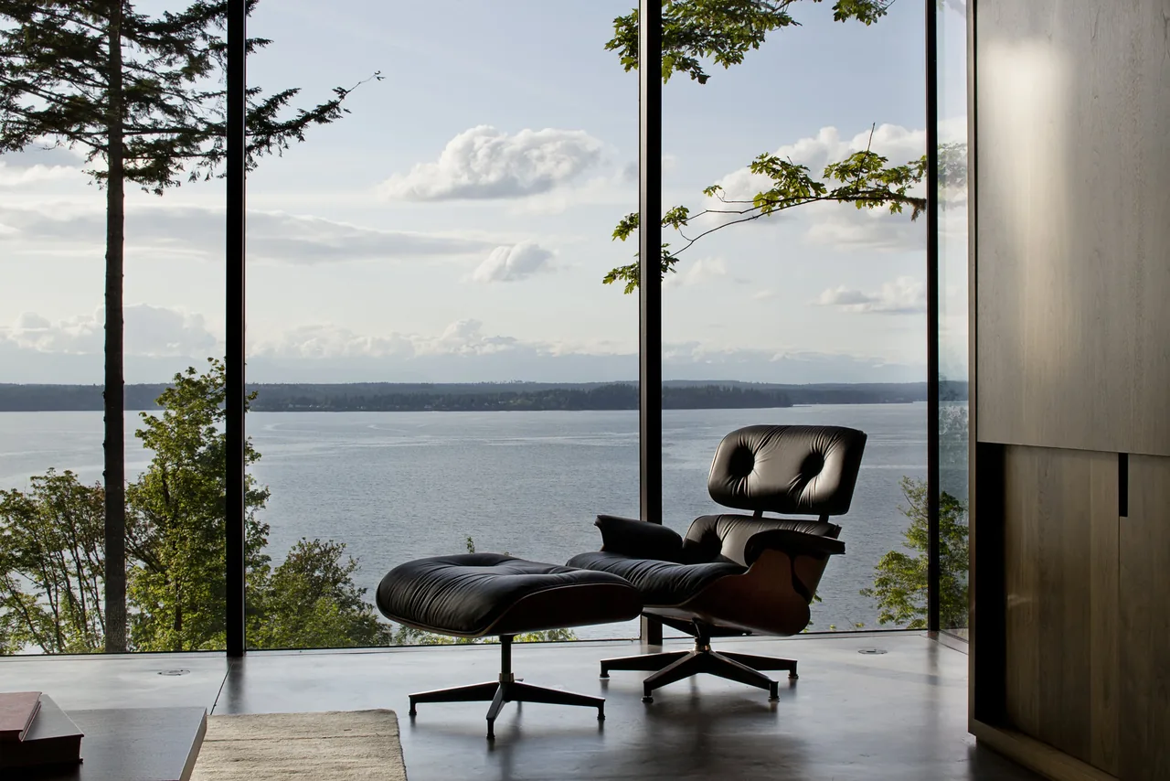 Black leather lounge chair in a room with floor-to-ceiling glazing that frames a lake and trees view.