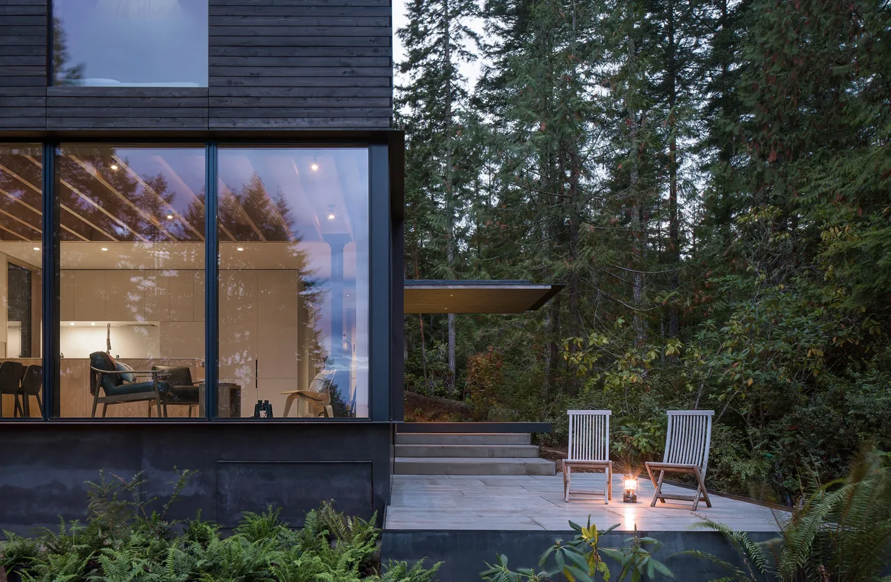 Magnolia residence features floor-to-ceiling windows and a patio with seating, set in a wooded landscape.