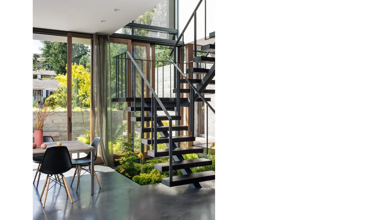 Dining area features a floating steel staircase and floor-to-ceiling wood-frame glass doors connecting indoor and outdoor spaces.