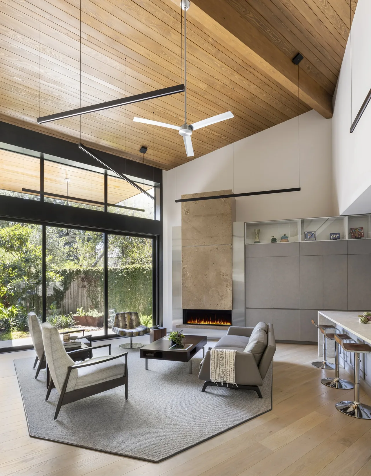 Shetland Lane House living room features a vaulted wood ceiling, large corner windows, and a linear fireplace with stone surround.