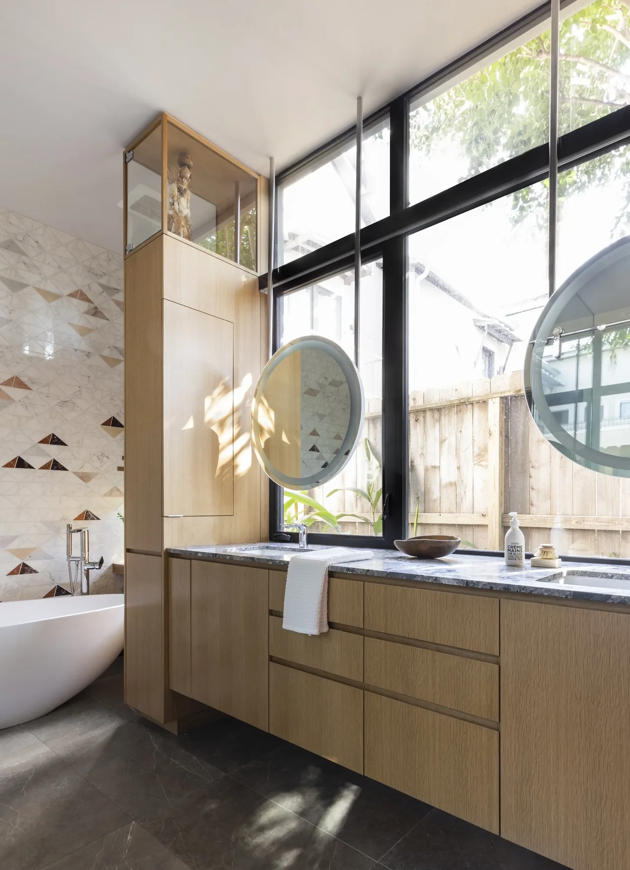 Bathroom with large window, wooden vanity, round mirrors, freestanding tub and geometric tile wall.