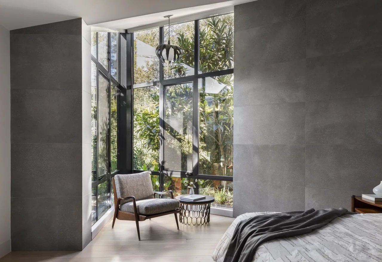 Bedroom corner features floor-to-ceiling black window frames with garden views and a wooden armchair by a round side table.