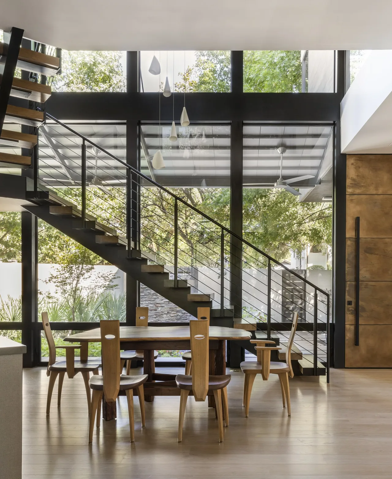 Dining space features floor-to-ceiling black-framed windows with a view of greenery and a metal staircase with wood steps.