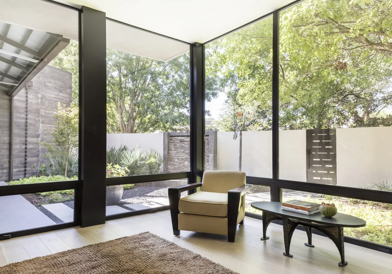 Living room corner features large floor-to-ceiling windows with black frames overlooking a landscaped garden.