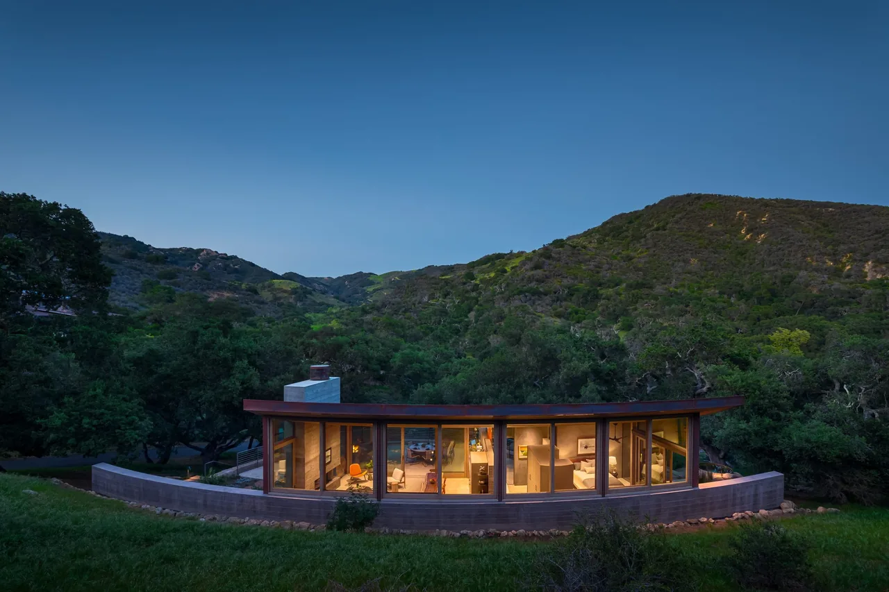 Curved guest house and studio with large windows nestles in a green hillside at dusk.