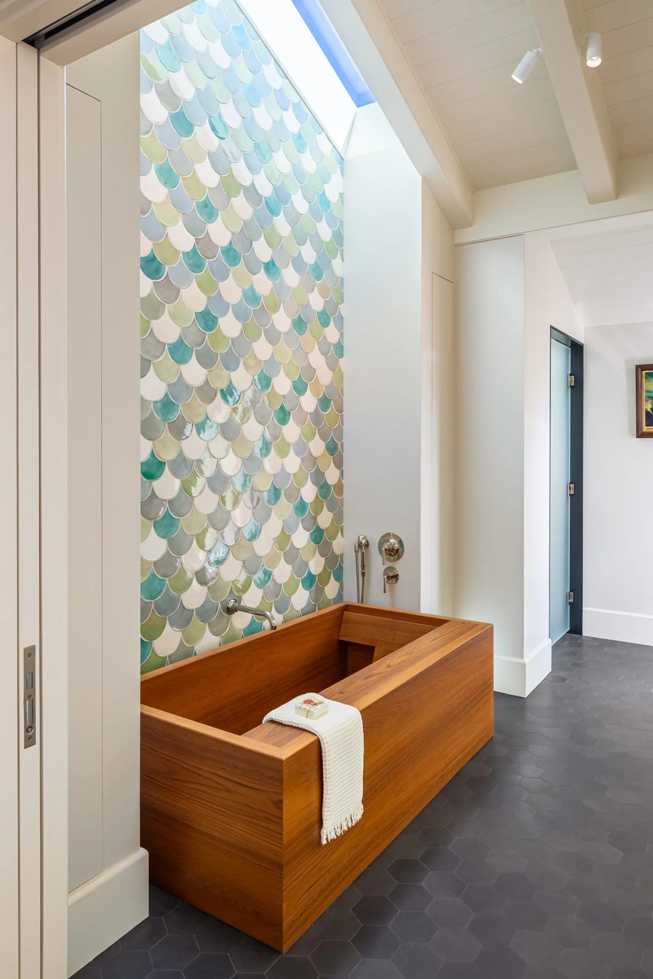 Bathroom features a rectangular wooden soaking tub with a multi-colored scalloped tile accent wall and natural light from a skylight.