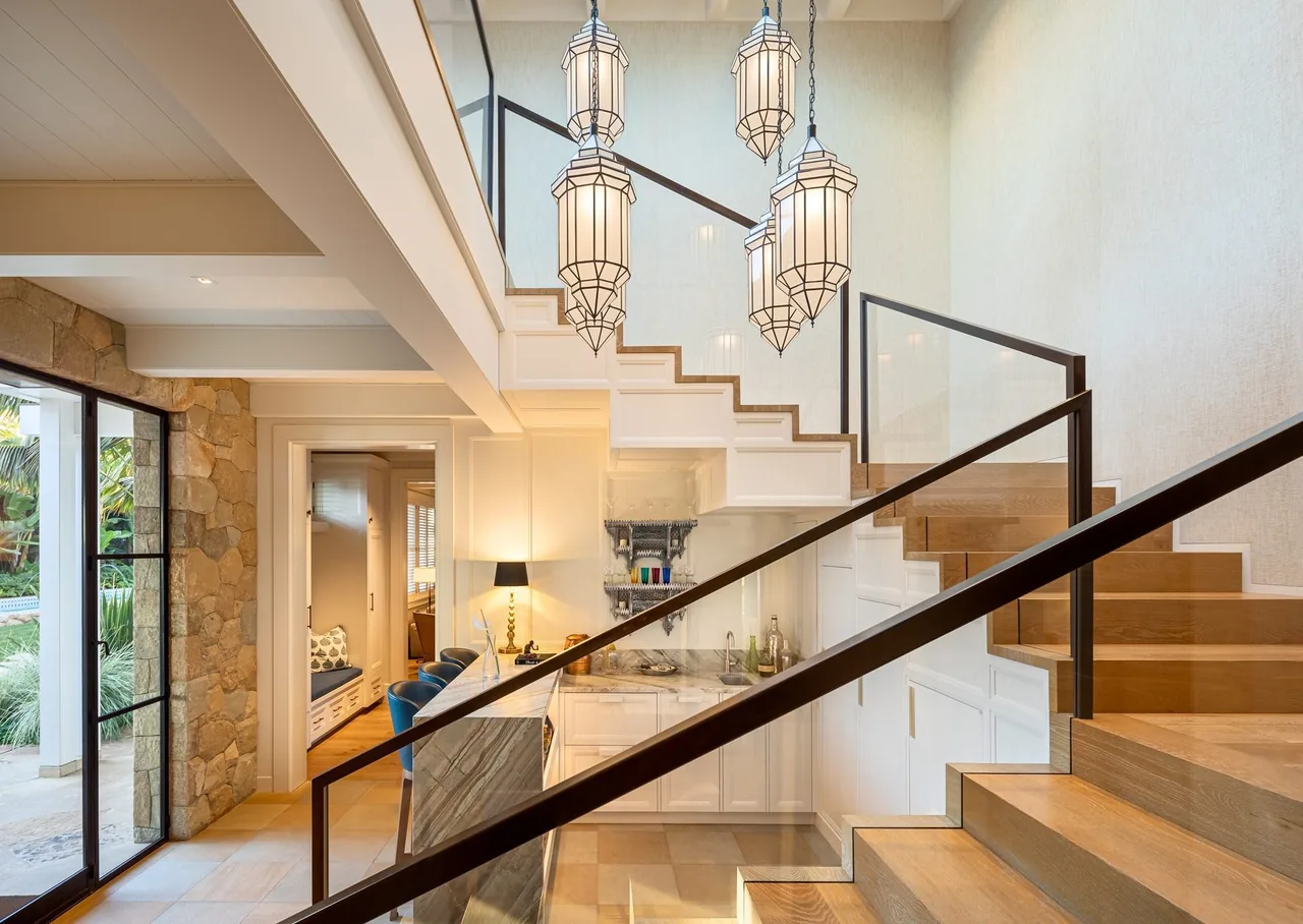 Staircase with glass railing and pendant lights overlooking a compact wet bar at Padaro Beach Home.