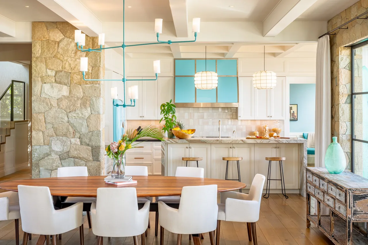 Indoor dining area featuring a wooden table and upholstered chairs next to a kitchen with blue accent cabinetry and a stone wall.