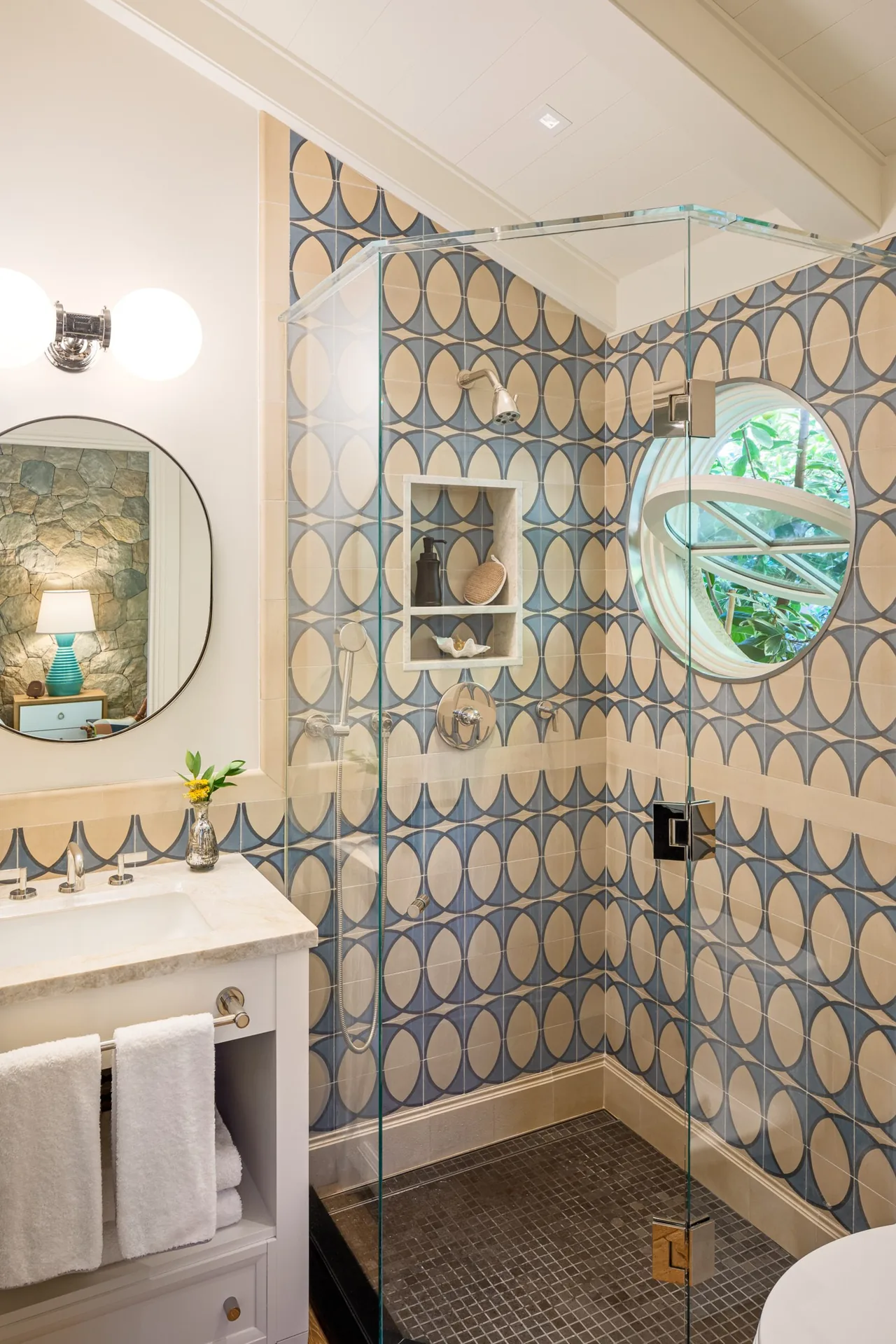 Shower area in a bathroom with scalloped patterned tiles and a round window with a garden view.