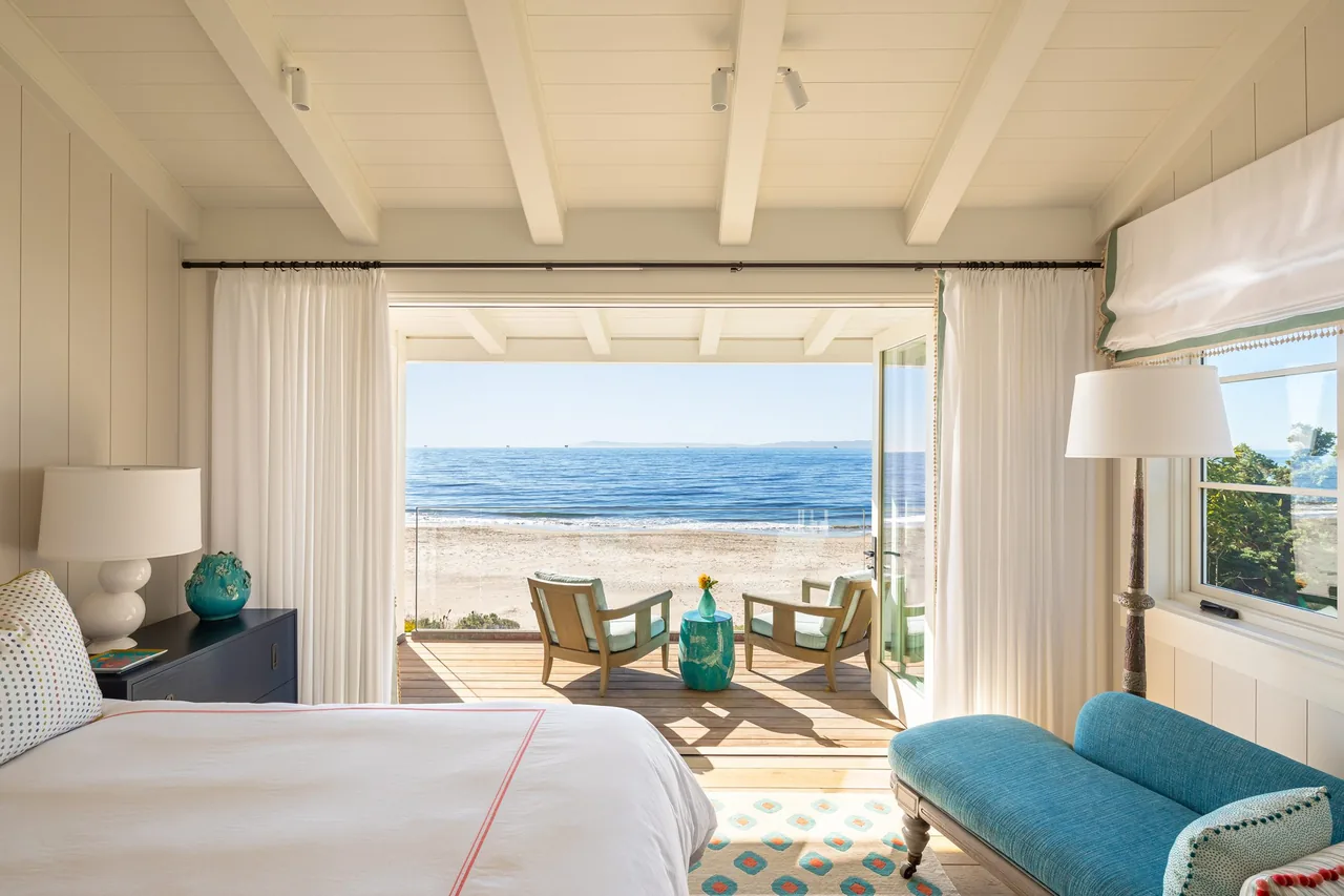 Bedroom with white bedding opens to a wooden deck with two chairs and ocean view at Padaro Beach Home.