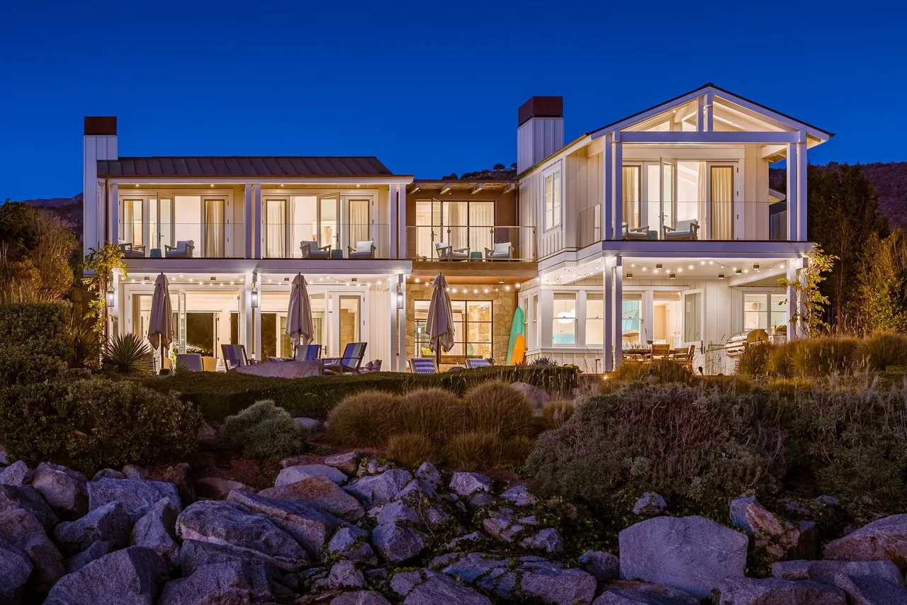 Padaro Beach Home features a two-story white exterior with balconies, extensive outdoor seating, and ambient string lighting at dusk.