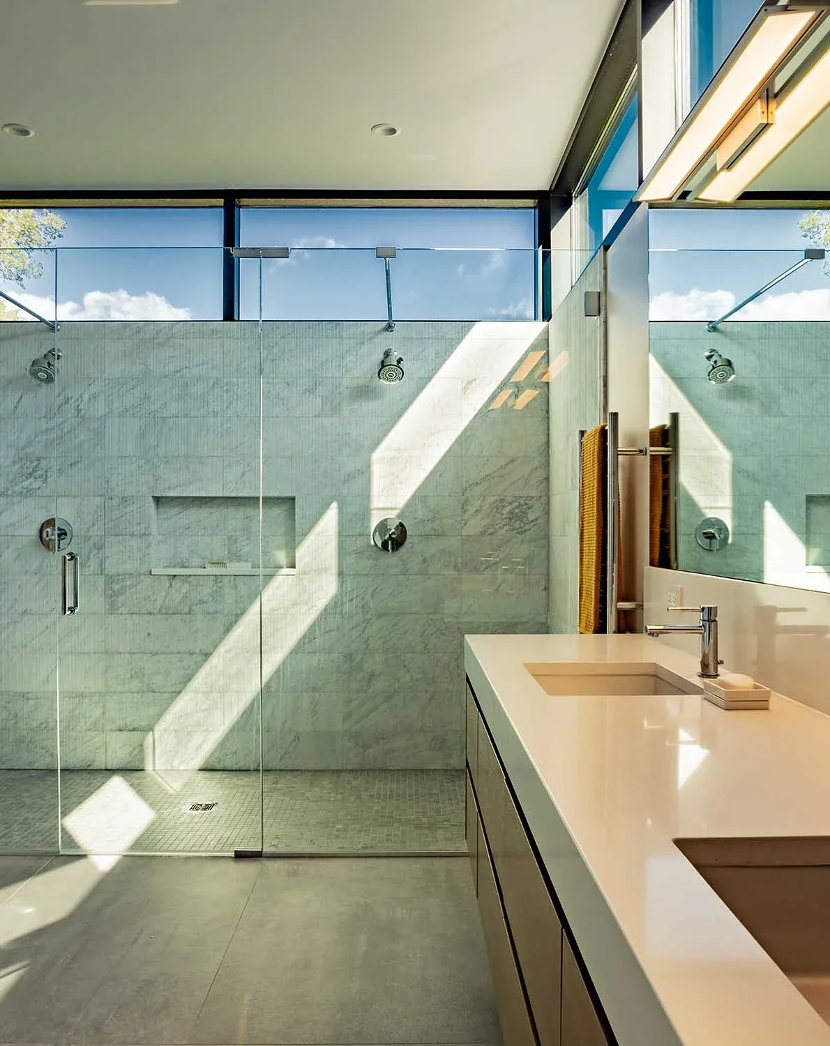 Modern bathroom with marble tile shower, glass door, and double vanity under natural light from clerestory windows.