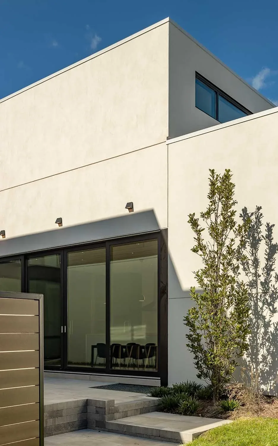 Modern two-story house exterior featuring black framed sliding glass doors and a small landscaped plant area.