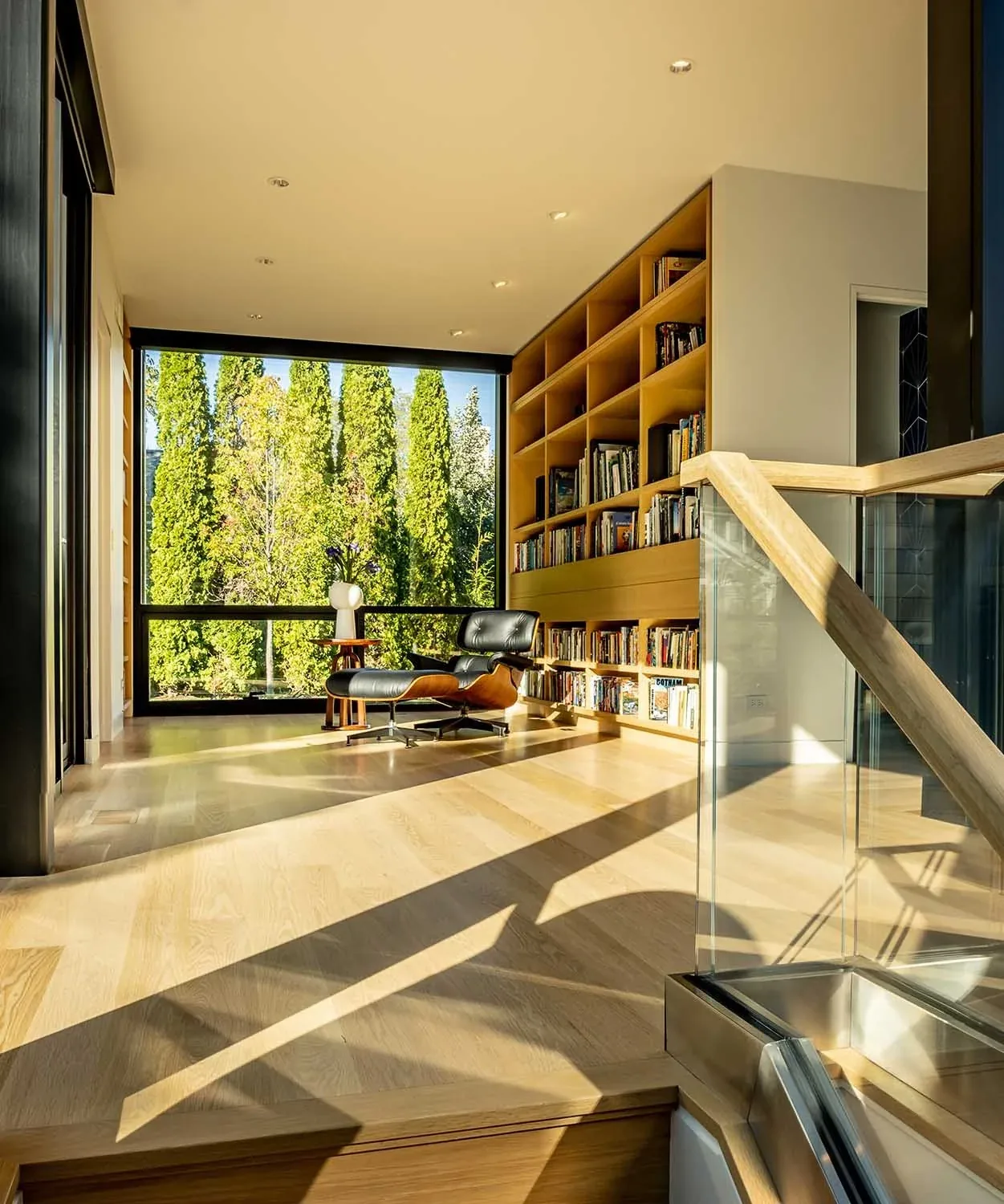 Upper-level reading nook featuring built-in wood bookshelves and a black leather lounge chair in front of a large window.