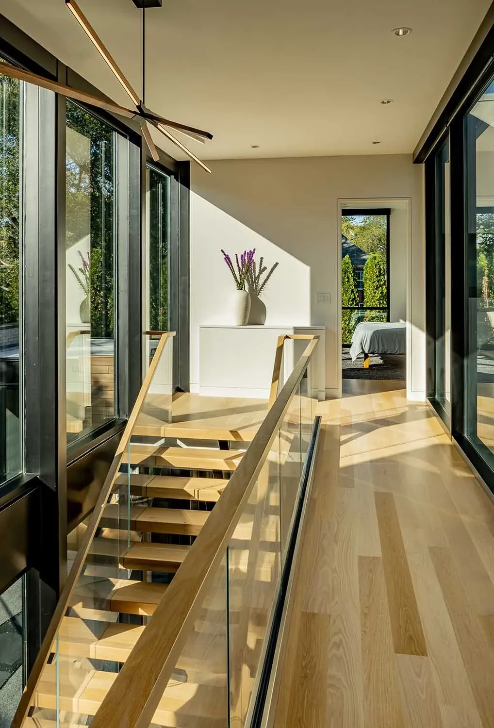 A bright hallway showcases floating wood stairs with a glass balustrade beside black-framed windows.