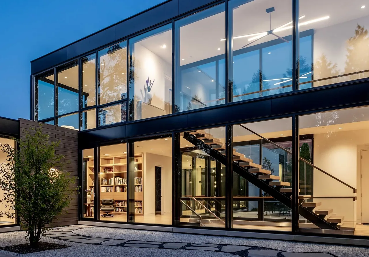 Two-story house with floor-to-ceiling glass walls and floating stairs visible through black-framed windows.