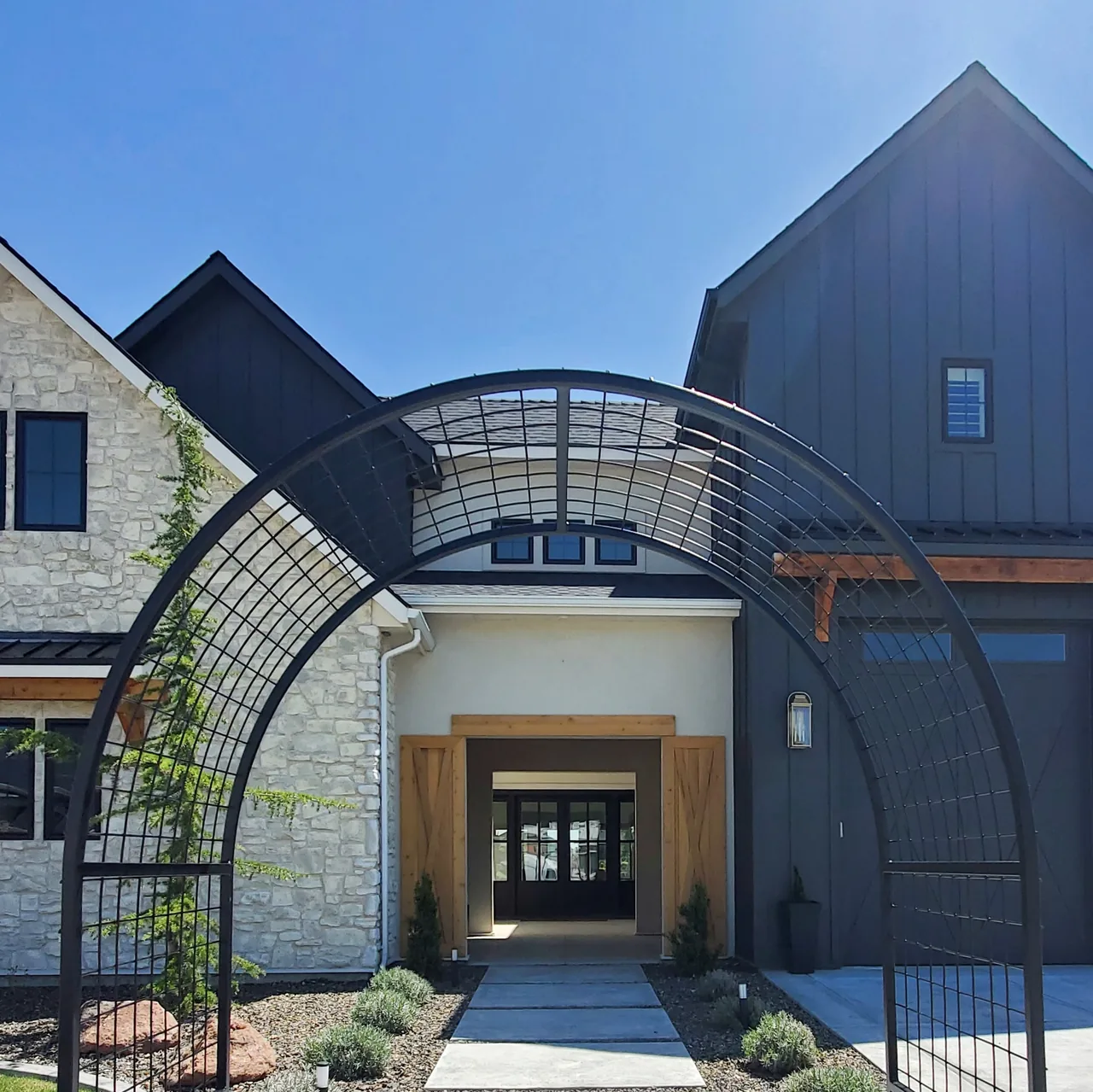 Front exterior of a house with mixed stone and dark siding, featuring a metal archway at the entrance pathway.