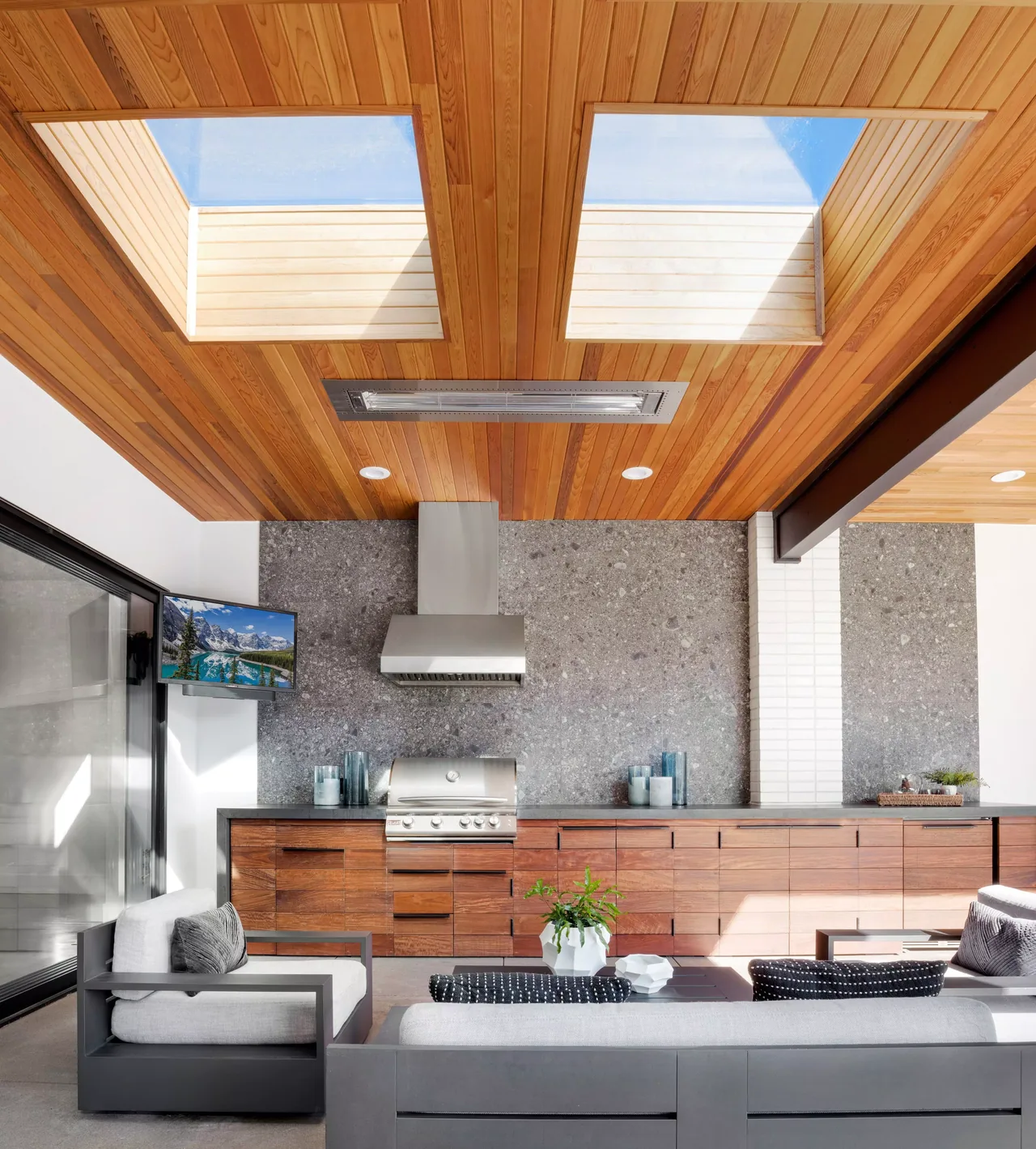 Outdoor kitchen and seating area feature wood cabinetry, a grill, stone backsplash, and skylights in the ceiling.