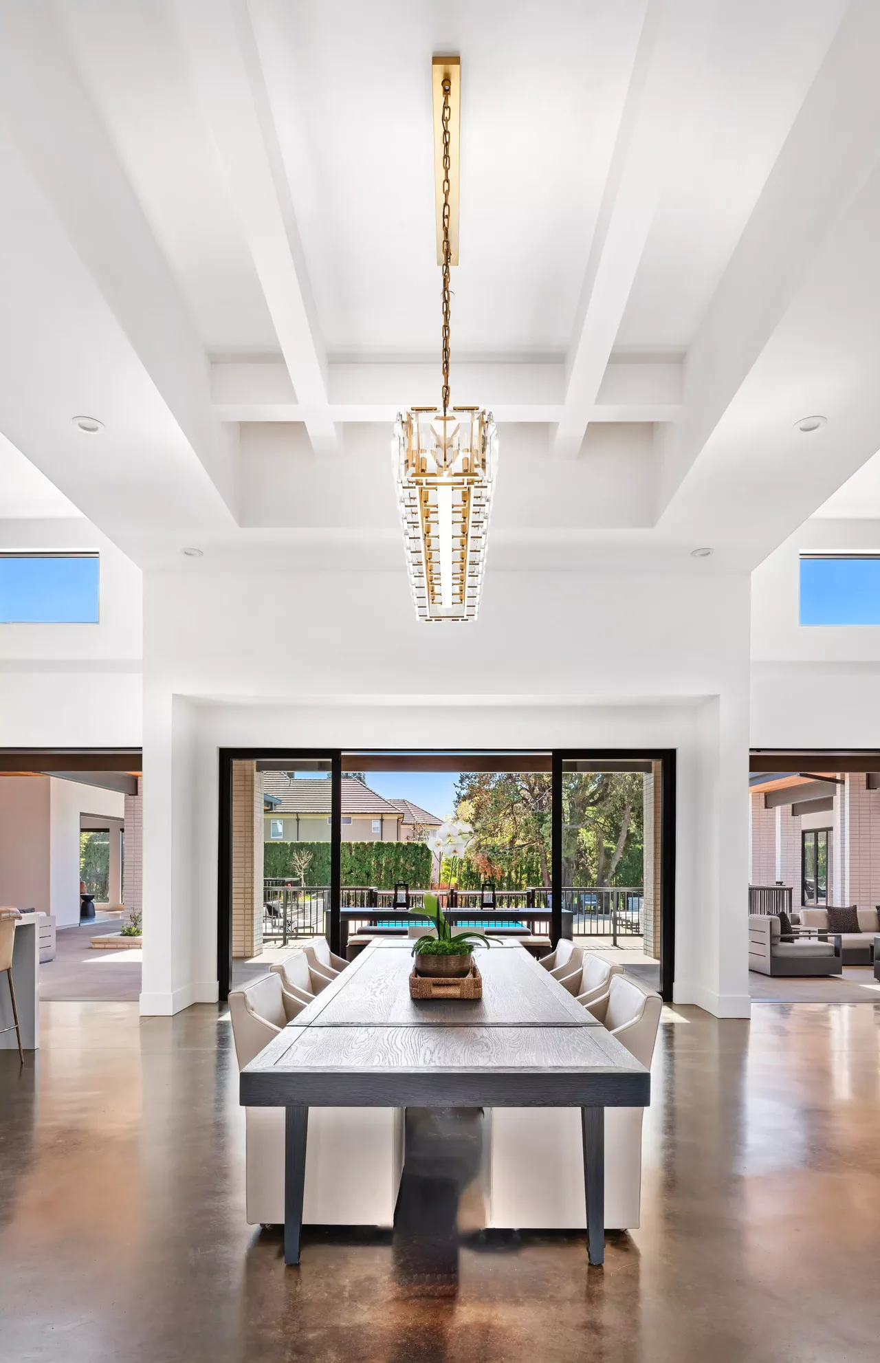 Dining room with a large wooden table and white chairs, featuring a geometric chandelier and floor-to-ceiling glass doors to an outdoor area.