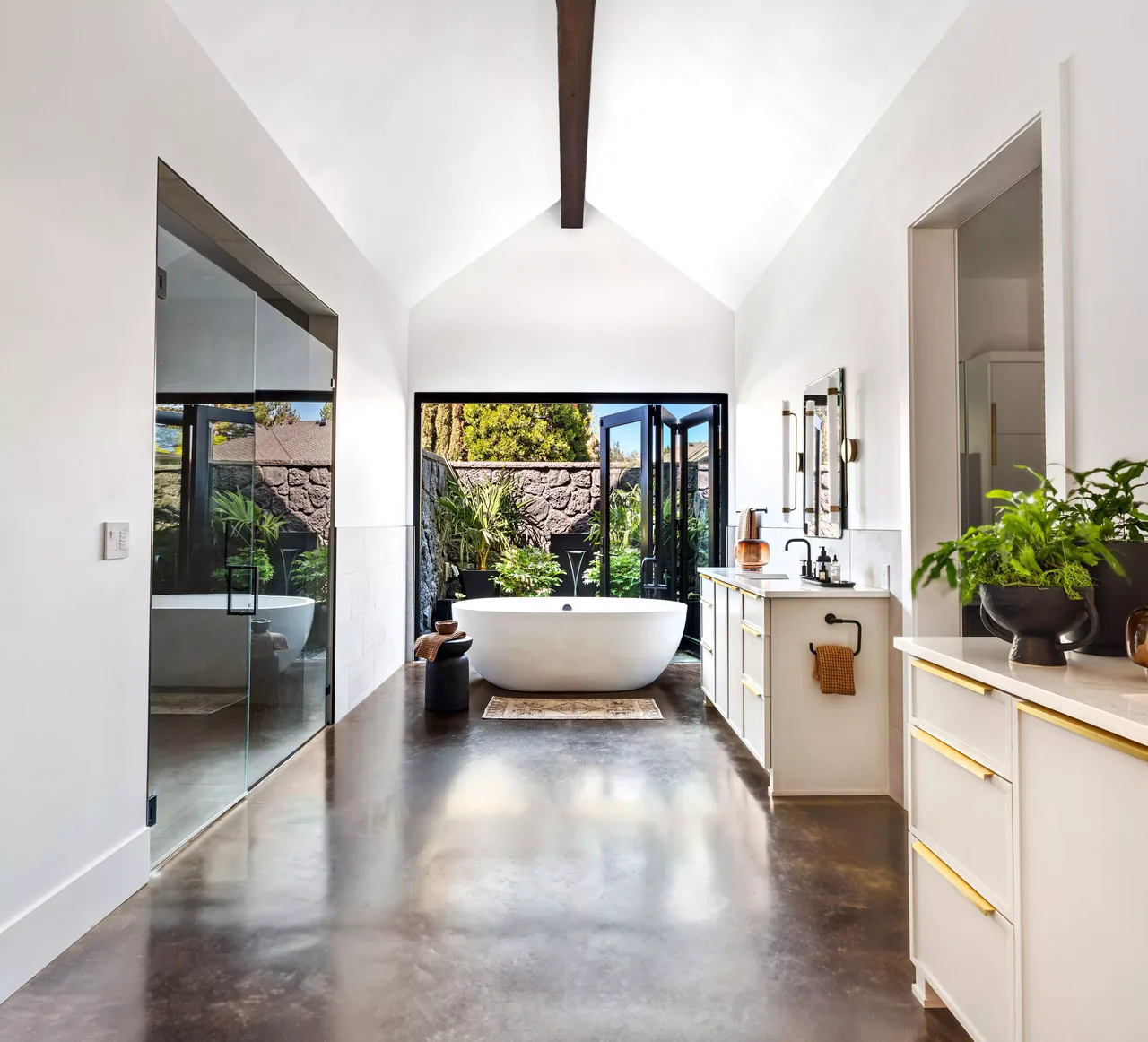 Modern bathroom with vaulted ceiling and floor-to-ceiling glass doors opening to a private garden area.