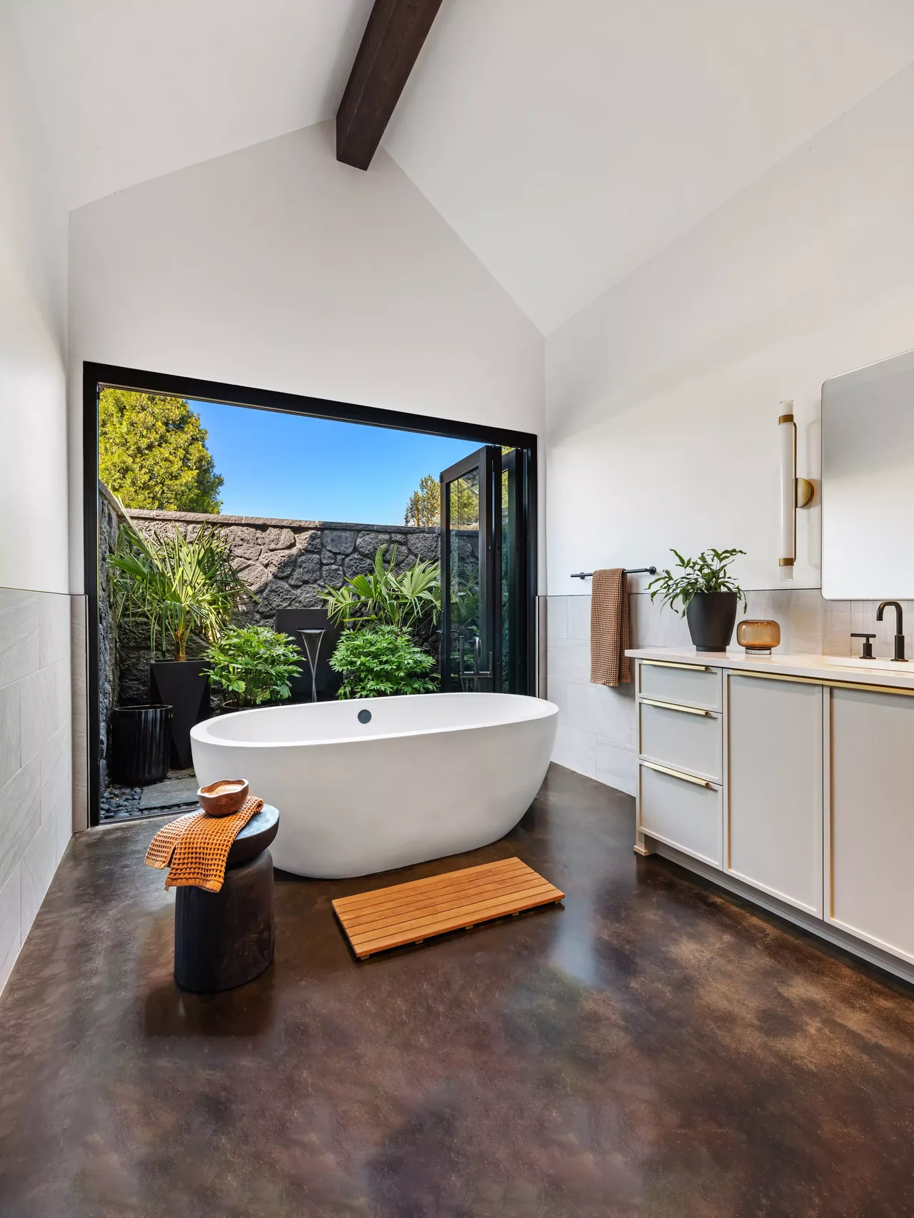 Modern bathroom featuring a freestanding tub with a large open window facing an outdoor garden walled with stone.