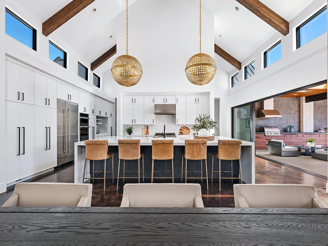 Modern kitchen with vaulted ceiling, wooden beams, white cabinetry, leather bar stools, and two round pendant lights, overlooking an outdoor seating area.