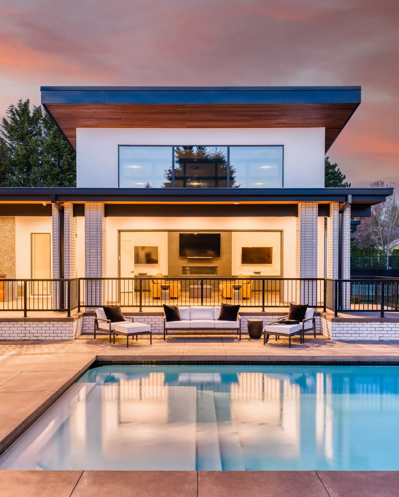 Modern home with black window frames and outdoor seating overlooking a pool with a pink and orange sky at dusk.