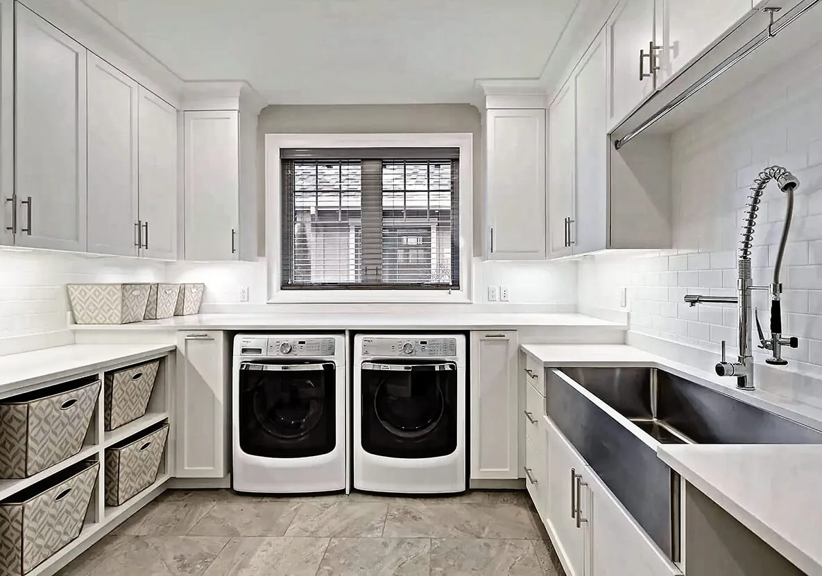 Laundry room features white cabinets, a large stainless steel sink with a flexible faucet, and front-loading washer and dryer under a window with blinds.
