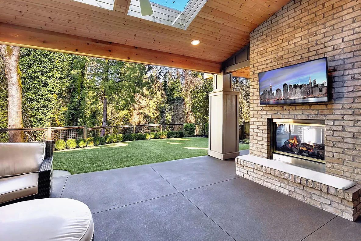 Covered patio featuring a brick fireplace with a mounted TV and a skylight above overlooks a fenced green lawn.
