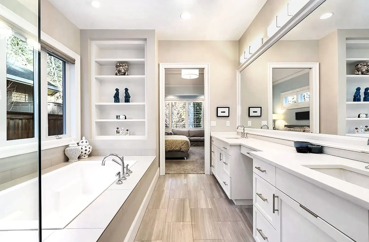 Modern bathroom with a built-in tub, double vanity, and large mirror, connecting to a bedroom with neutral tones.