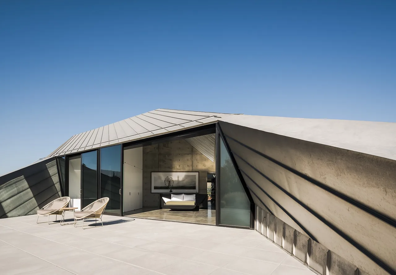 Exterior view of a modern building with a geometric sloped roof and an open glass door leading to a bedroom.