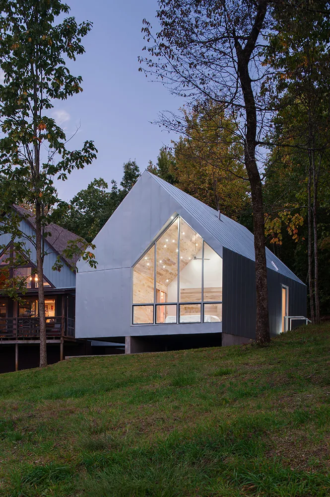 A modern cabin with a steep gable roof and large floor-to-ceiling windows set on a grassy hillside surrounded by trees.
