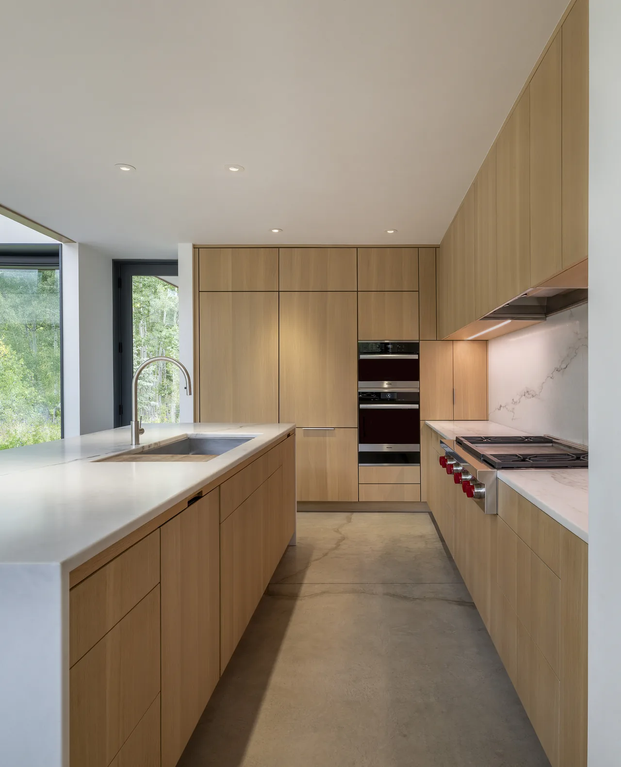 Kitchen with floor-to-ceiling light wood cabinets, marble countertops, and large windows overlooking greenery.