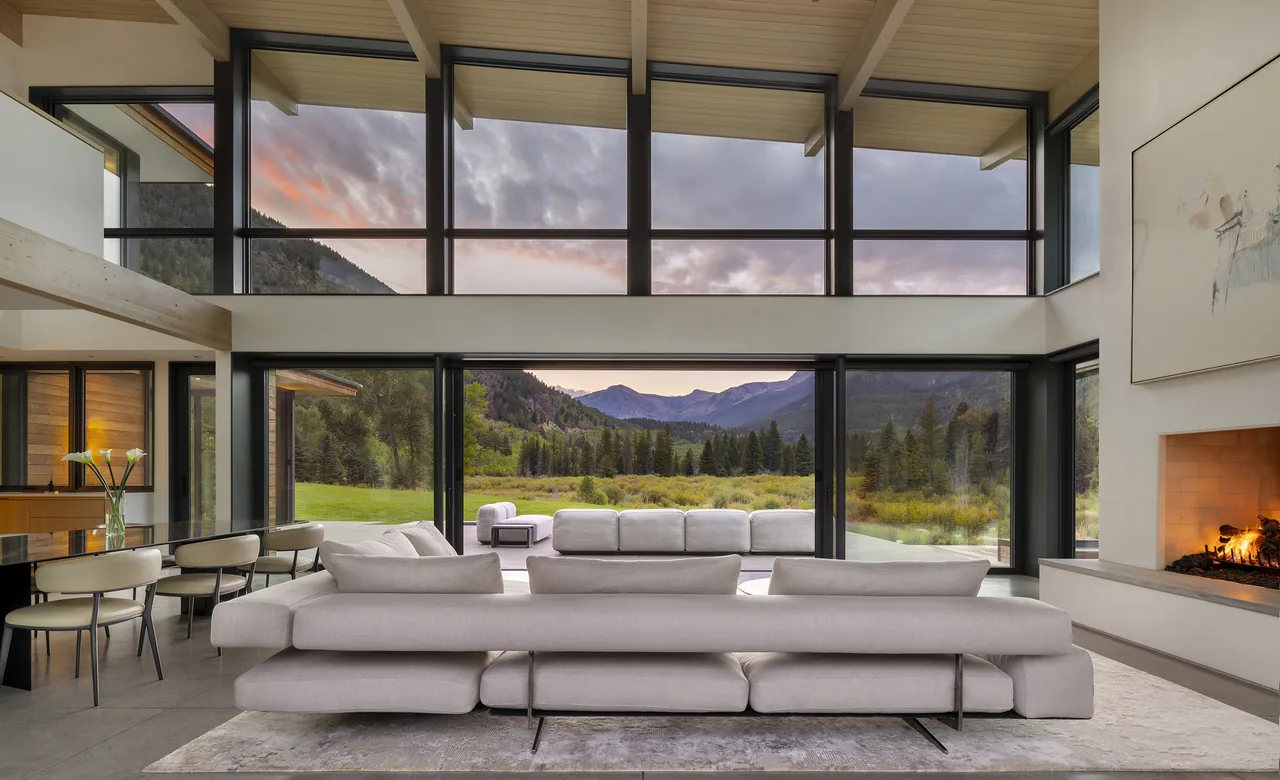 Living room with floor-to-ceiling black-framed windows, a fireplace, and a view of mountains and greenery at dusk.
