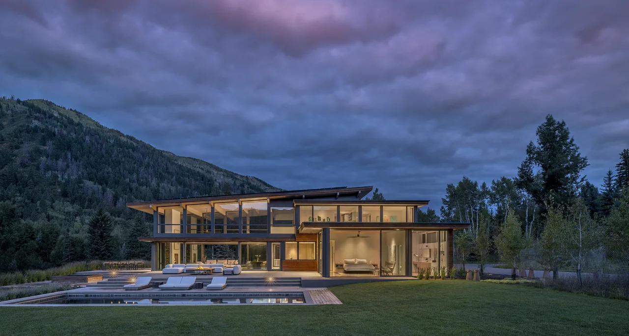 Modern two-story home with floor-to-ceiling glazing and poolside chaise lounges overlooking a mountain backdrop.