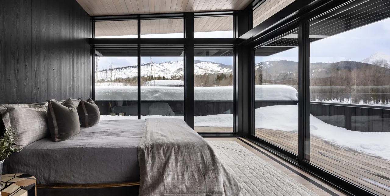 Bedroom with black window frames and floor-to-ceiling glazing overlooks a snow-covered deck and mountain scenery.