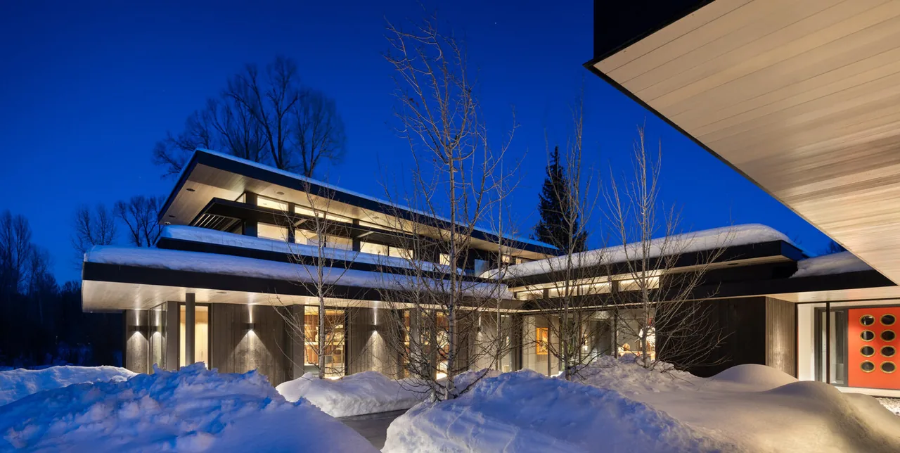Nighttime image of a modern home with flat roofs covered in snow and warm interior lighting in a snowy landscape.