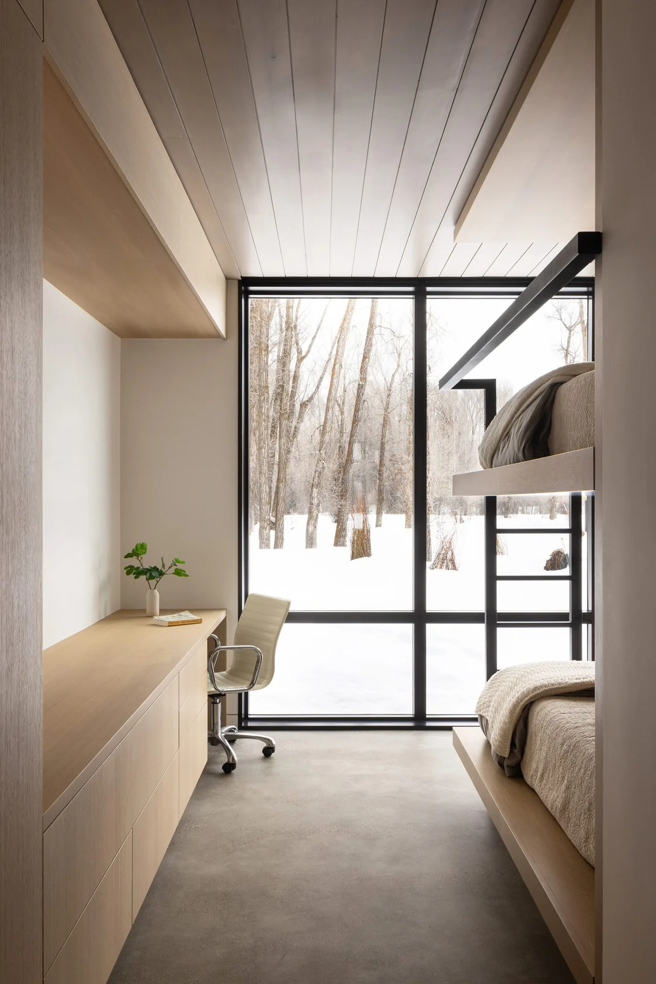 Light wood bunk beds and built-in desk stand beside large floor-to-ceiling black-framed window with snowy view.