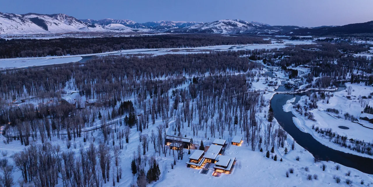 Thunder Road Reaches Jackson Hole, Wyoming, a contemporary home glowing with warm lights amid snow and bare trees at dusk by a river.
