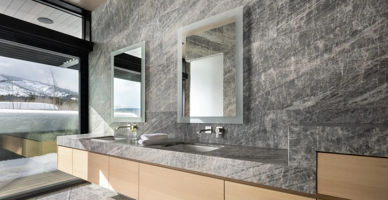 Bathroom vanity with two mirrors and sinks, featuring striking stone surfaces and a large window overlooking snow-covered mountains.