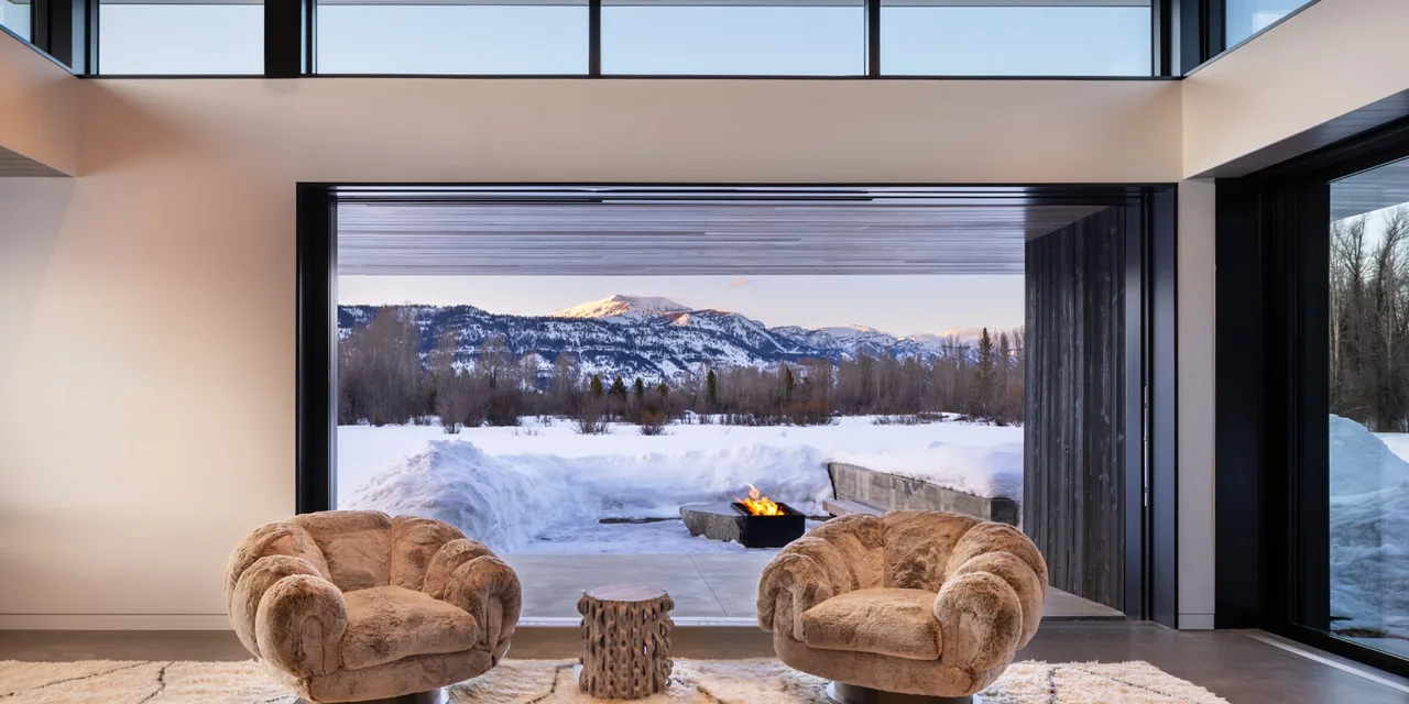 Living room interior with two plush armchairs and large retractable glass doors opening to a snowy mountain view and outdoor fire pit.