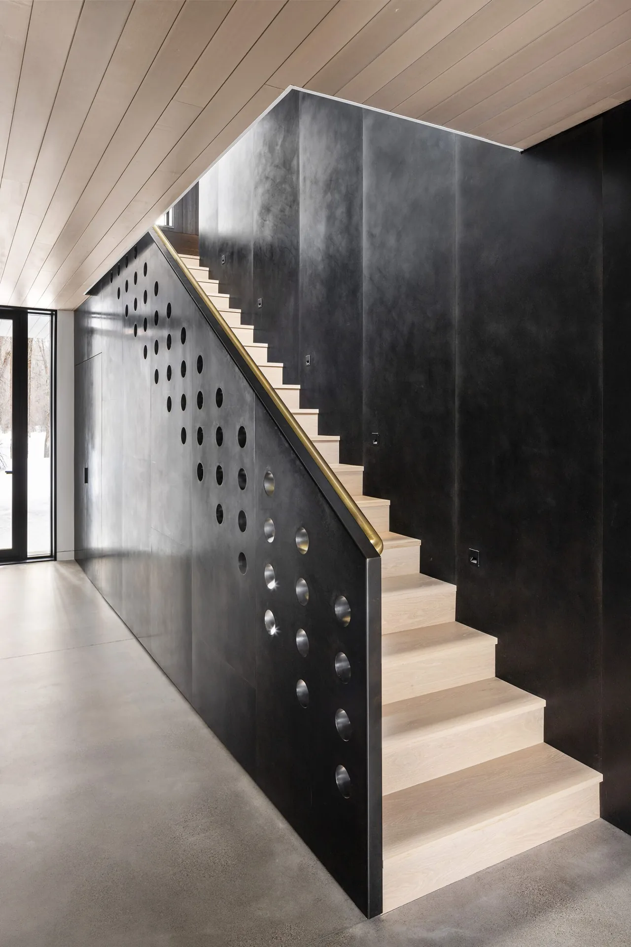 A modern staircase with light wood treads and perforated black panel railing in a sleek interior.