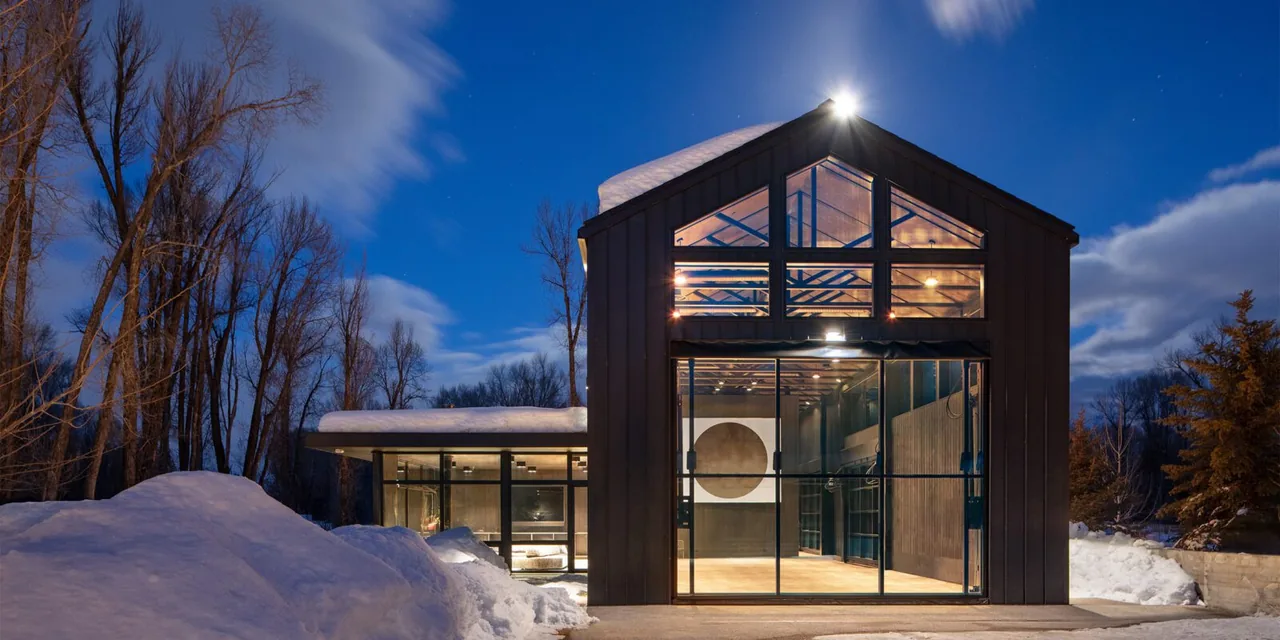 The modern home features floor-to-ceiling black window frames and a snow-covered gable roof at twilight.