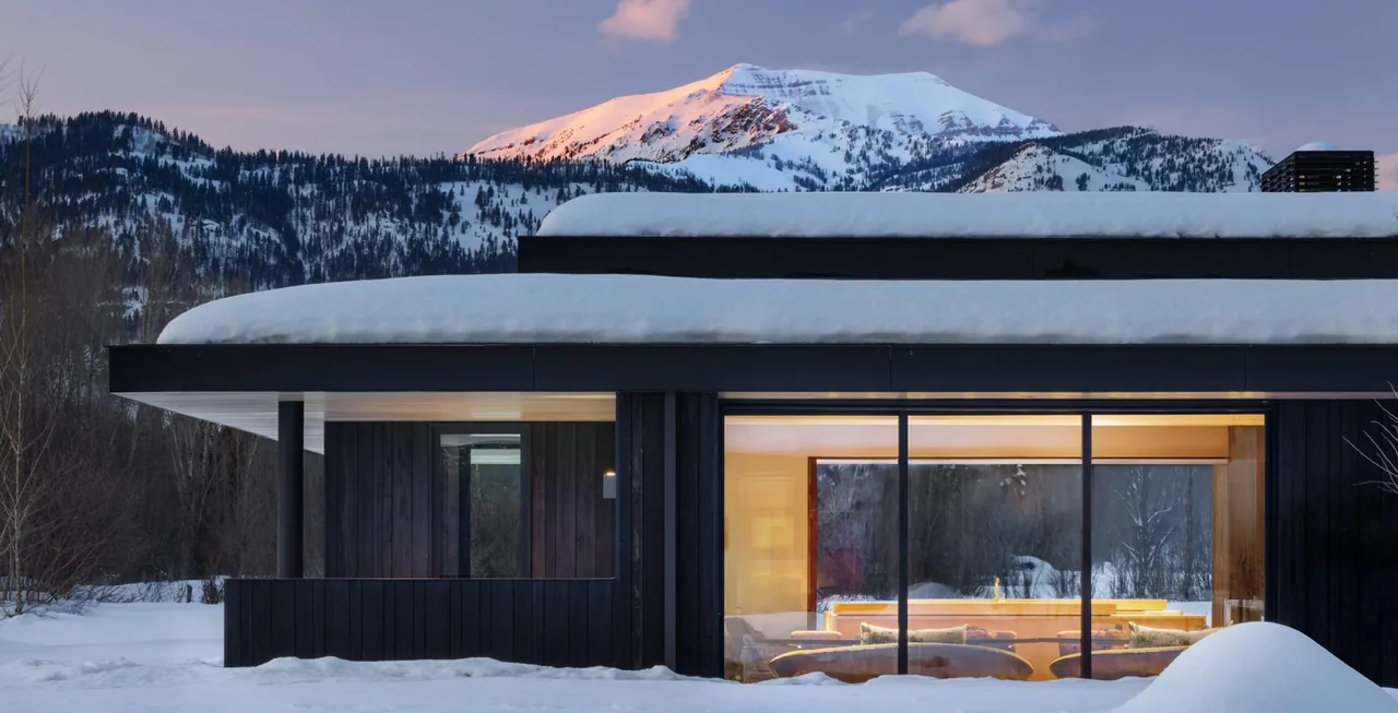 The Thunder Road house features a flat roof and expansive glass walls with snowy mountain views at twilight in Jackson Hole, Wyoming.