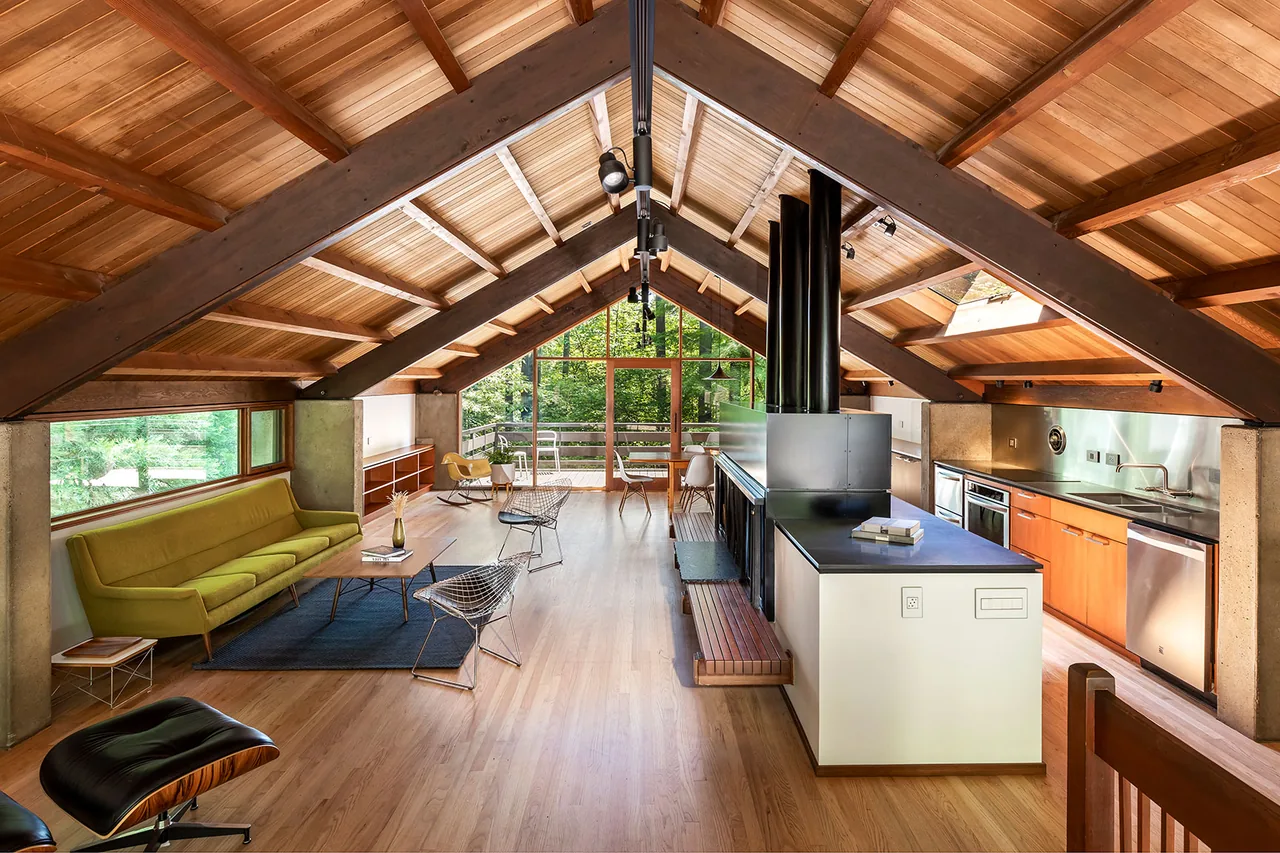 Open living and kitchen area under a vaulted wood-beamed ceiling with large windows and a central fireplace.
