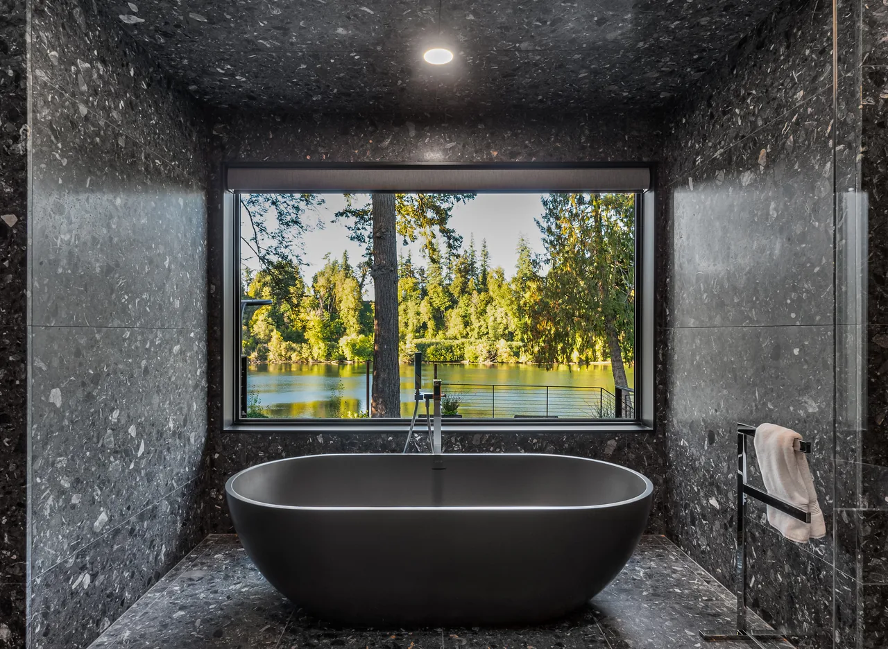Bathroom with dark stone walls and freestanding black bathtub positioned below a large picture window with a forest and lake view.